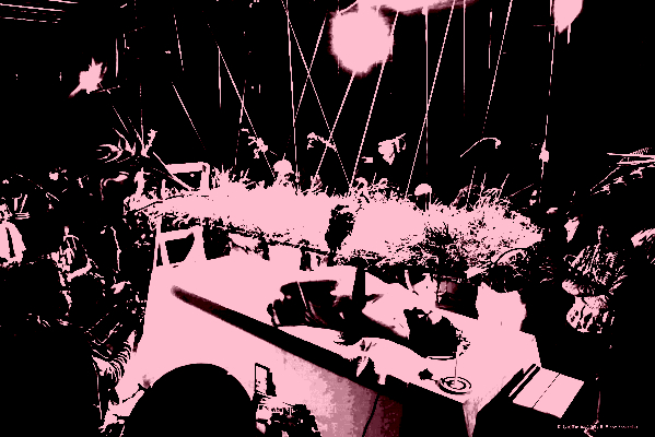 This image depicts a person lying on a table in the middle of a crowd holding a plane of bushy grass, plants and flowers that is suspended horizontally above them.