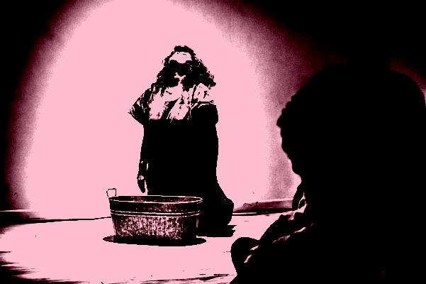 This image depicts a person with long, curly black hair is kneeling, looking upwards towards the light, perhaps imploring the heavens. The baggy fitting red dress stands out vividly against the background, and contrasts with the cold hard metal tub set in front of her.