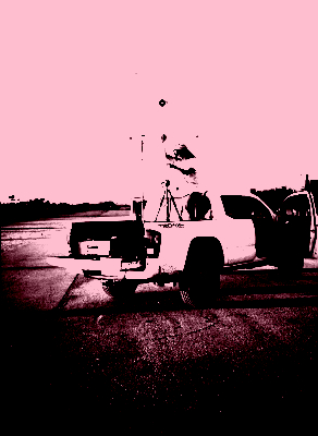 This image depicts a person standing in the back of a white pick-up truck In an empty asphalt lot, installing radio antennas resembling crosses on tripods. This curious setup seems to pick up sounds and transmit them to a computer.