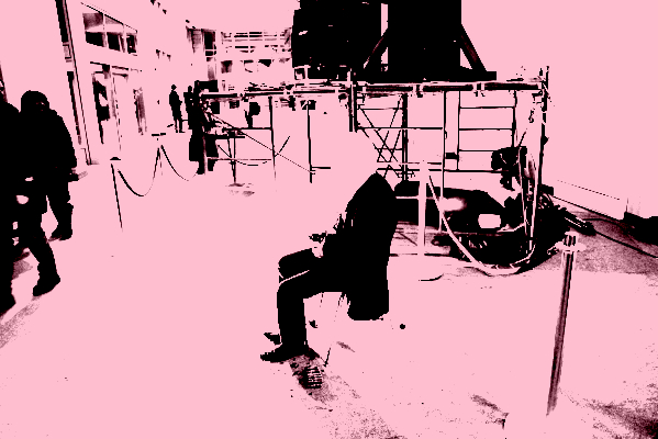 This image depicts a person sitting on a chair amid some debris cutting pennies with a pair of pliers. His head is covered by a pillowcase, and red velvet crowd-control barriers for galas are placed around him.
