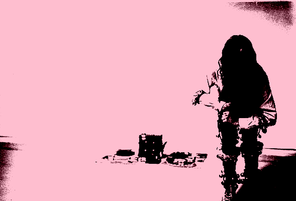 This black-and-white analogue photograph depicts a person standing crouched in front of a mat covered in percussion instruments and music equipment. Bells adorn their elbows, knees and ankles.